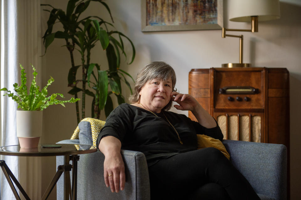 I don't know who this person is. A woman sits in an armchair in a living room with a radio, plant, and a side table with a potted plant next to her