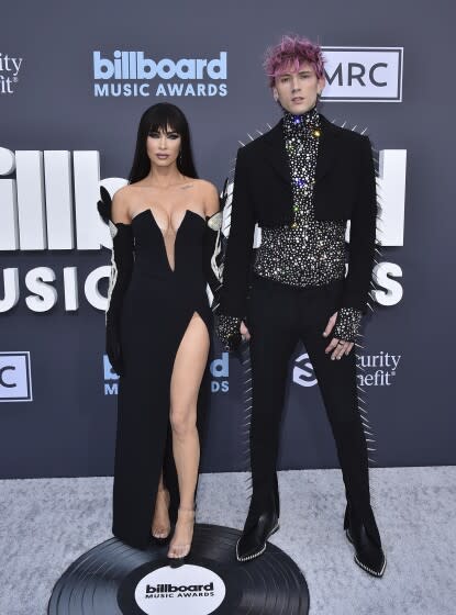 A woman in a black gown and a man in a spikey black suit pose for cameras on a gray carpet