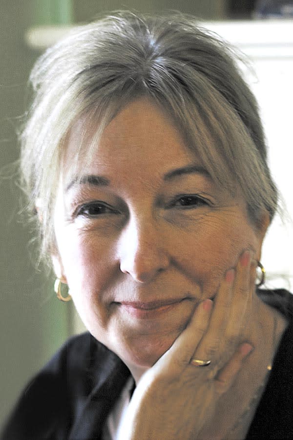 A gray-haired white woman wears gold hoops, rests her chin in her hand, and smiles slightly to the camera.