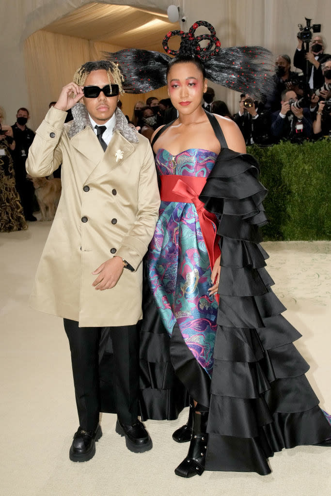 Naomi Osaka wears a floor length gown with a brightly colored bodice and Cordae Dunston wears dark slacks and a trench coat