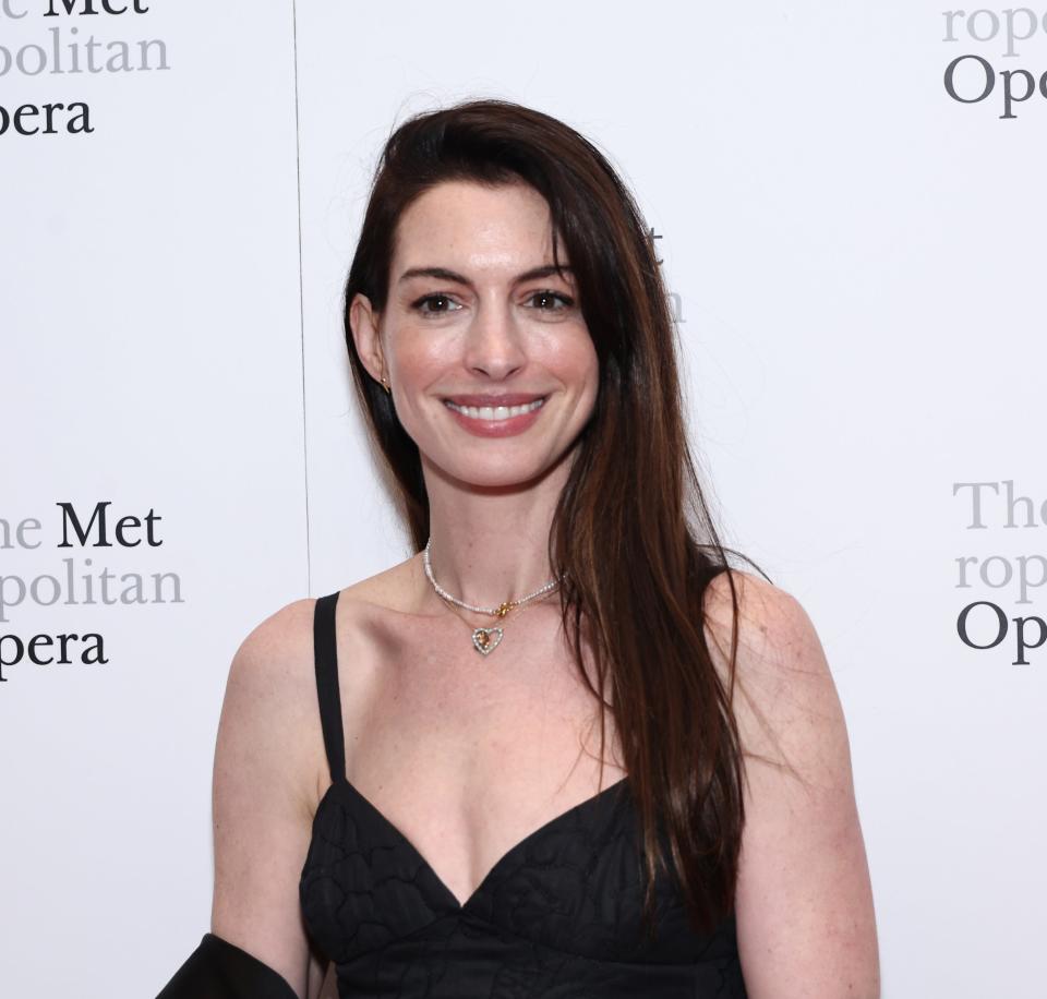 Anne Hathaway attends The Metropolitan Opera event, wearing a black dress with thin straps and a gold necklace. Smiling while posing for the camera