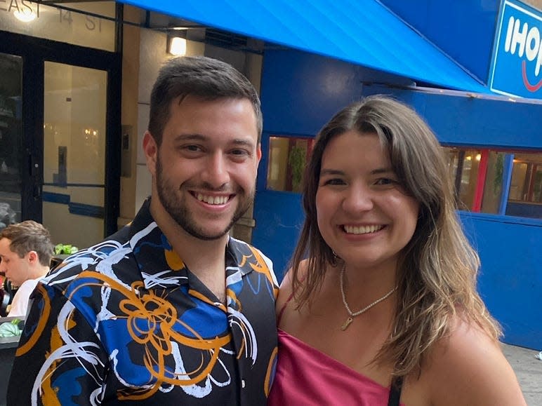 Meredith Wilshere and her boyfriend smiling and staring at the camera, standing outside on a sidewalk in front of a blue awning.