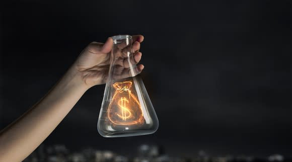 A hand holding up an Erlenmeyer flask containing a glowing image of a bag marked with a dollar sign.