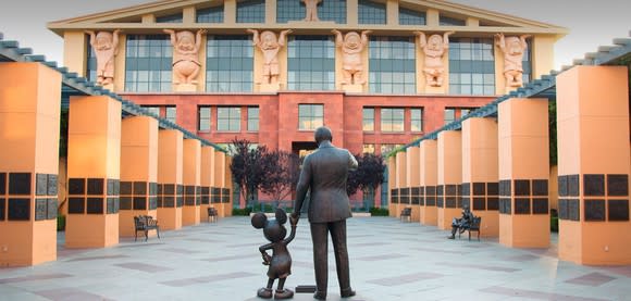 A statue of Walt Disney and Mickey Mouse holding hands in the middle of a path to the entrance of a Disney building.