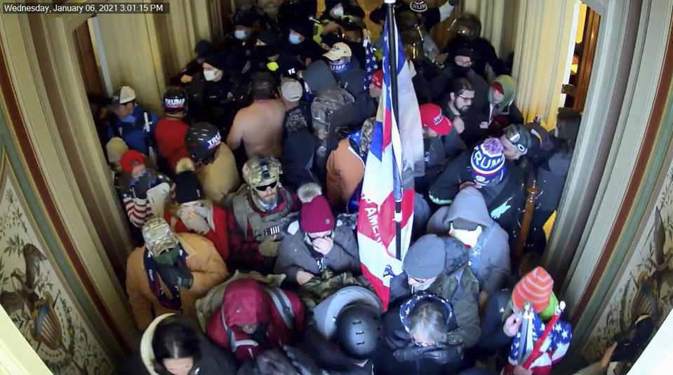 This image provided by the FBI taken from a video security camera in the U.S. Capitol building on Jan. 6, 2021, shows a man wearing a Trump 2020 winter cap and a bullet proof vest beside a wooden coat rack, standing among other rioters. The FBI identified the man as Dustin Thompson. Federal authorities say Thompson illegally entered the U.S. Capitol on Jan. 6 and while he was there stole the coat rack. (The FBI via AP)