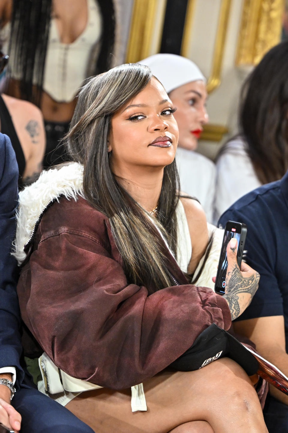 Rihanna attends a fashion show wearing a casual outfit with an oversized jacket and holding a smartphone