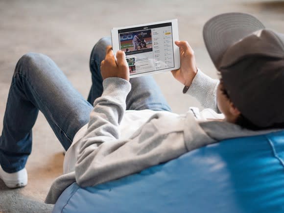 A young man with a ball cap sitting on a beanbag chair watching ESPN on an iPad.