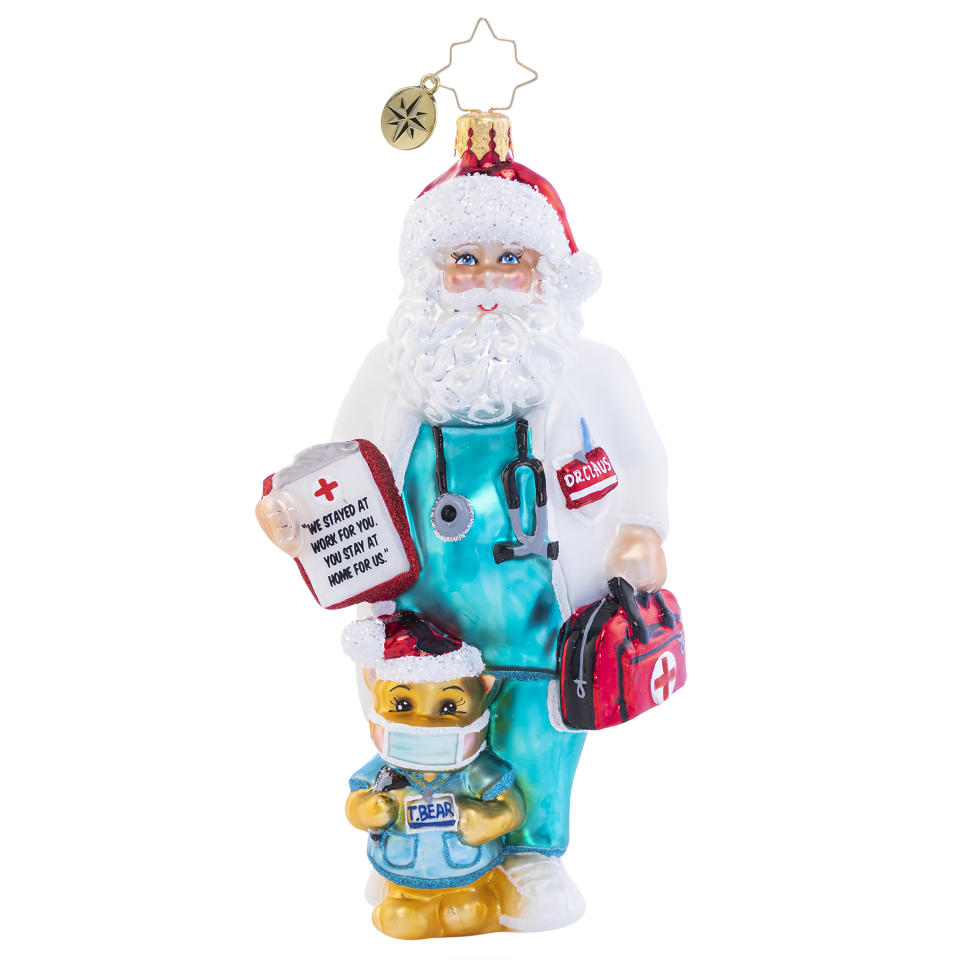 This image shows a Christopher Radko holiday ornament of a Santa in scrubs holding a message: “We stayed at work for you. You stay at home for us.” (Christopher Radko via AP)