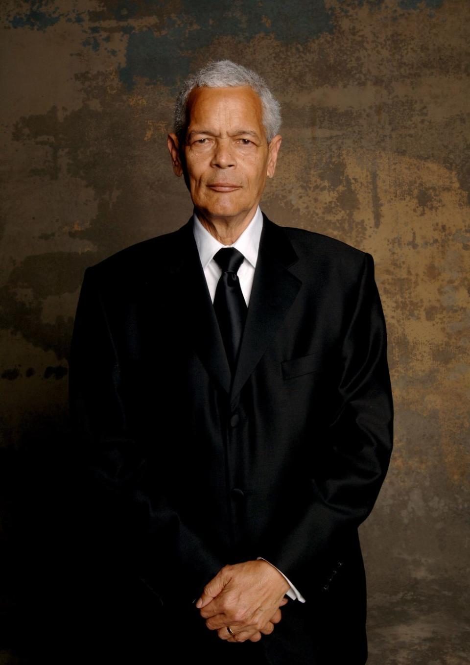 julian bond stands and looks at the camera with his hands clasped in front of him, he wears a black suit and tie with a white collared shirt