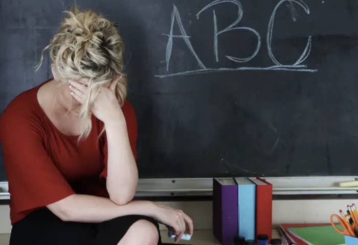 A stressed teacher sitting at a desk with her head in her hands by a blackboard with "ABC" written on it
