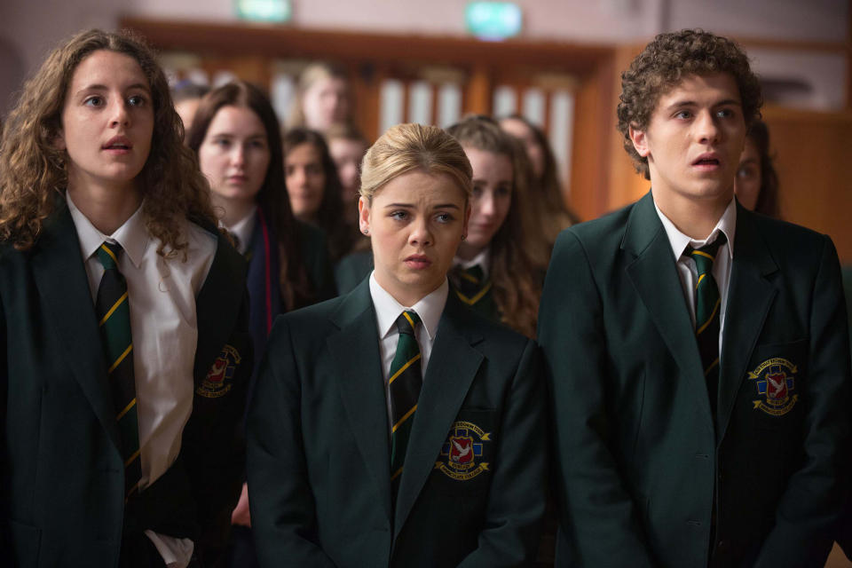 Three students stand at a school assembly. They all wear uniforms.