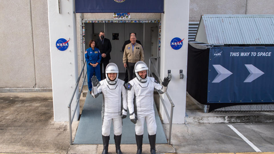 Astronauten der Besatzung 9 in weißen und schwarzen Raumanzügen salutieren, während sie sich auf den Weg zu ihrer Startrampe machen.