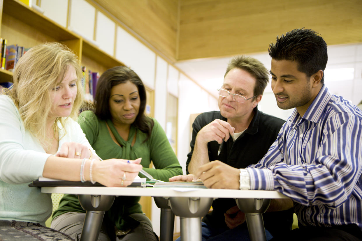 Candid photograph of a diverse group of mature students working together on a class project in their college library.  The students are sitting around a table in their college library concentrating hard while looking down at their project work on the desk in front of them.