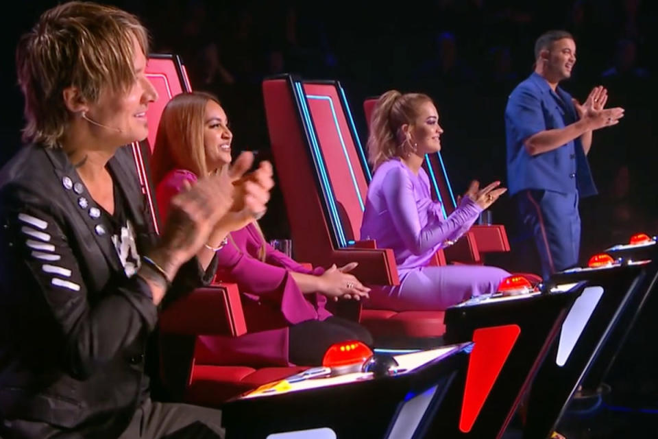 Guy Sebastian is in a blue shirt, standing and clapping. Rita Ora dressed in purple, Jessica Mauboy dressed in pink and Keith Urban in a black vest are all sitting on their The Voice chairs while clapping.