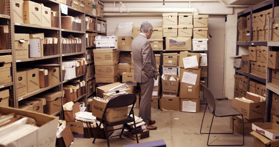A man in a gray suit stands in a room lined with boxes.