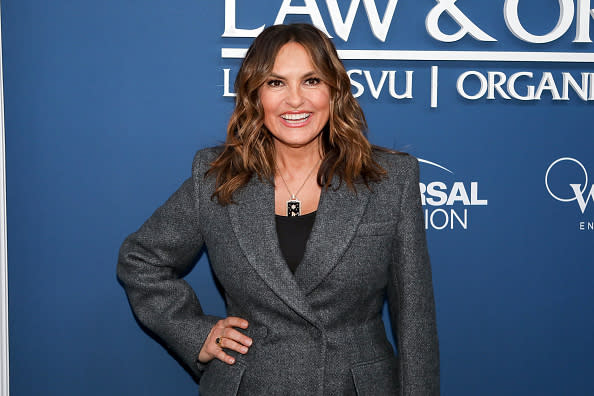 Mariska Hargitay smiles on the red carpet, wearing a stylish grey blazer, in front of a blue backdrop with "Law & Order" logos