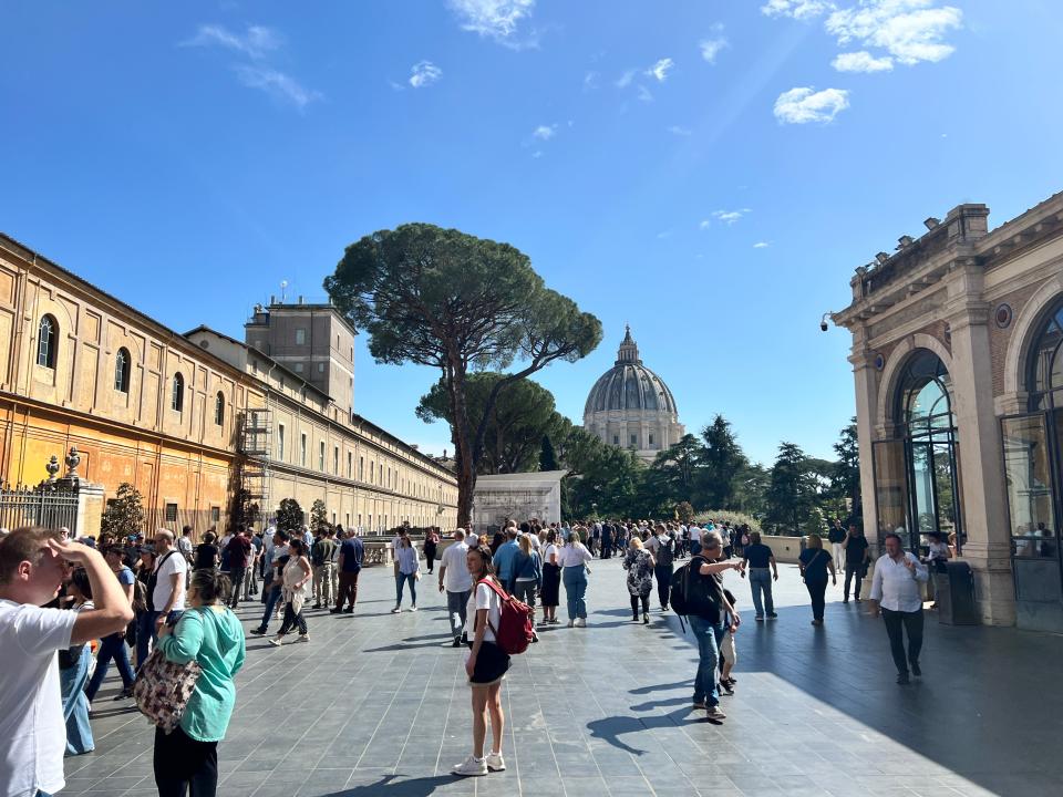 Tourists walking on the terrace of the Vatican Museums on a sunny day. St Peter's Basilica is visible in the background.