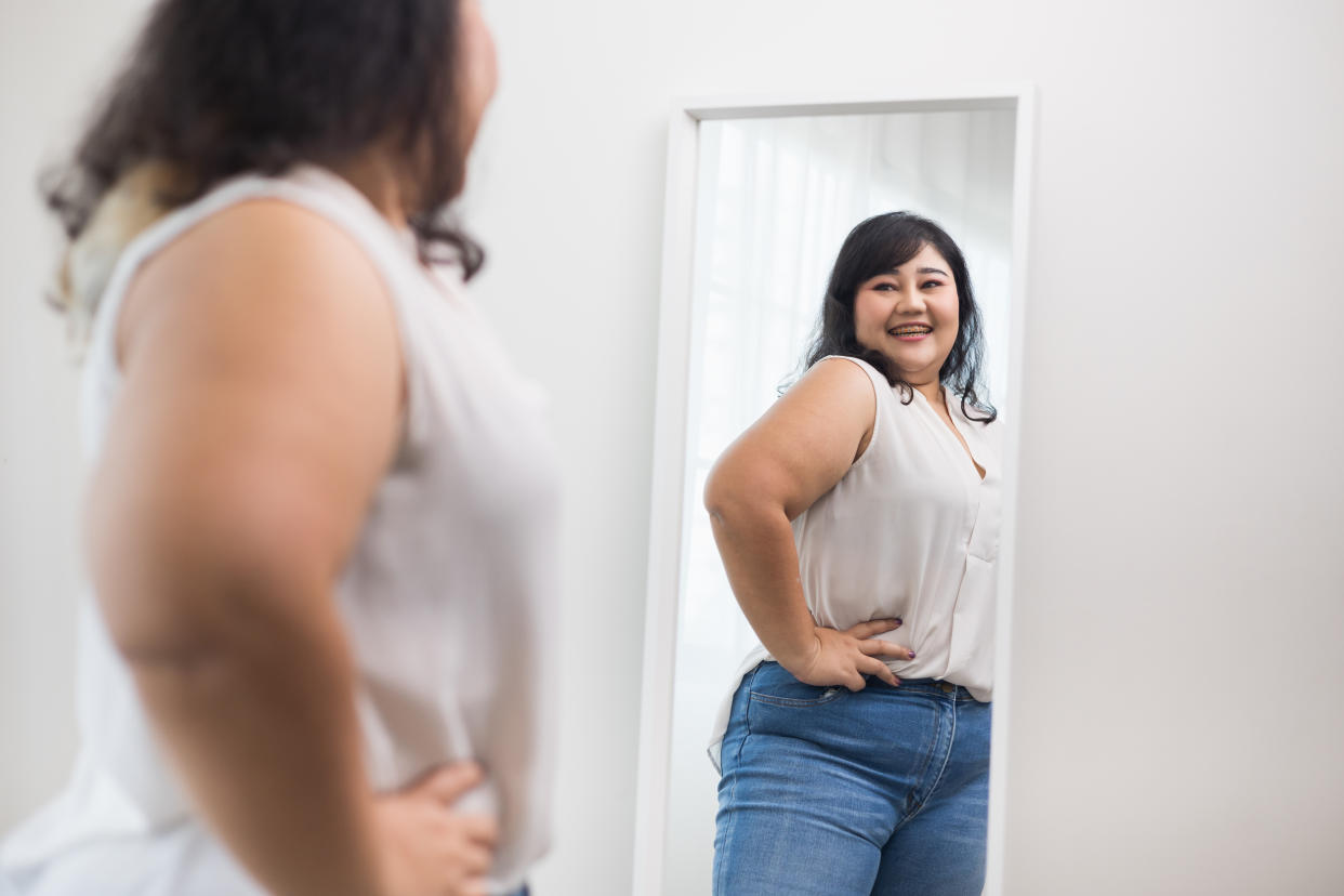 A woman wearing jeans and a white top looks into a full-length mirror.