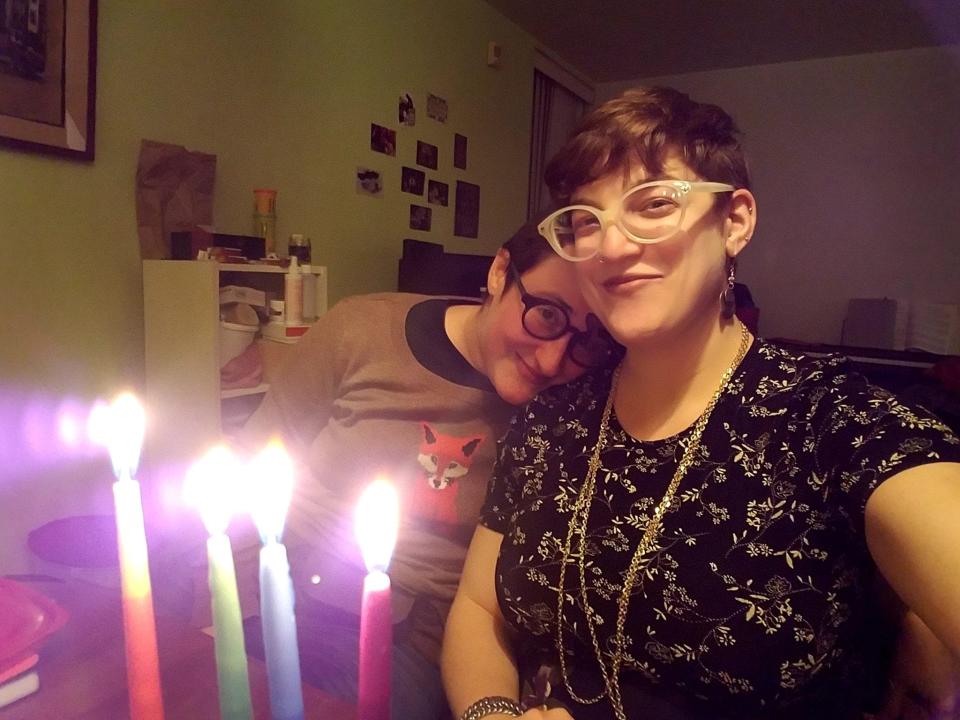 Two people pose for a selfie with a lit menorah, one resting their head on the other's shoulder.