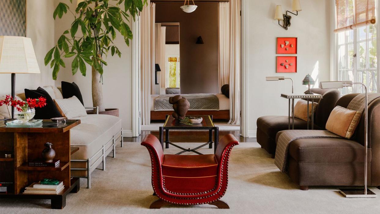 a living room with facing sofas, cocktail table with a sculpture atop between them, small red leather sleigh chair, large potted plant, floor lamps by sofa, end table with shelves, sconce above artworks, bamboo shades