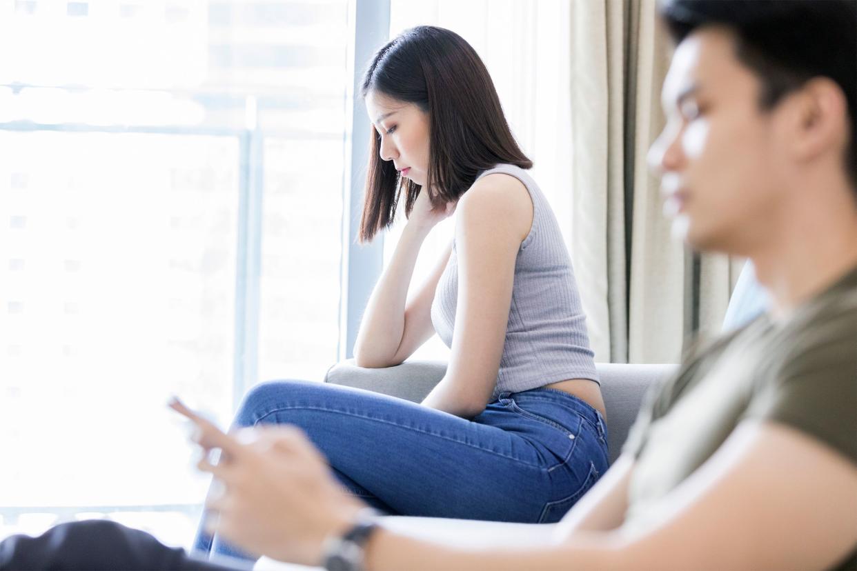 Woman and man sitting on opposite side of couch away from each other, woman in view, distraught, men is in foreground, blurred, window of high-rise apartment with sunlight coming in, in the background