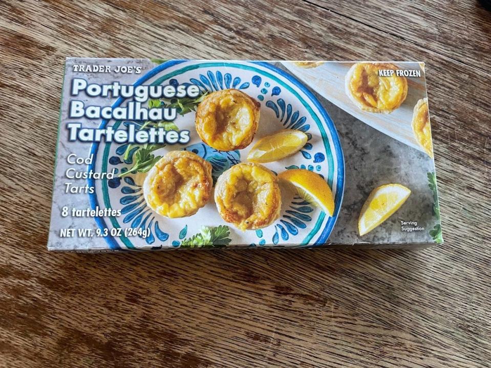 Box of Trader Joe's Portuguese Bacalhau tartelettes with a picture of tarts and blue plate on the packaging on a wooden counter