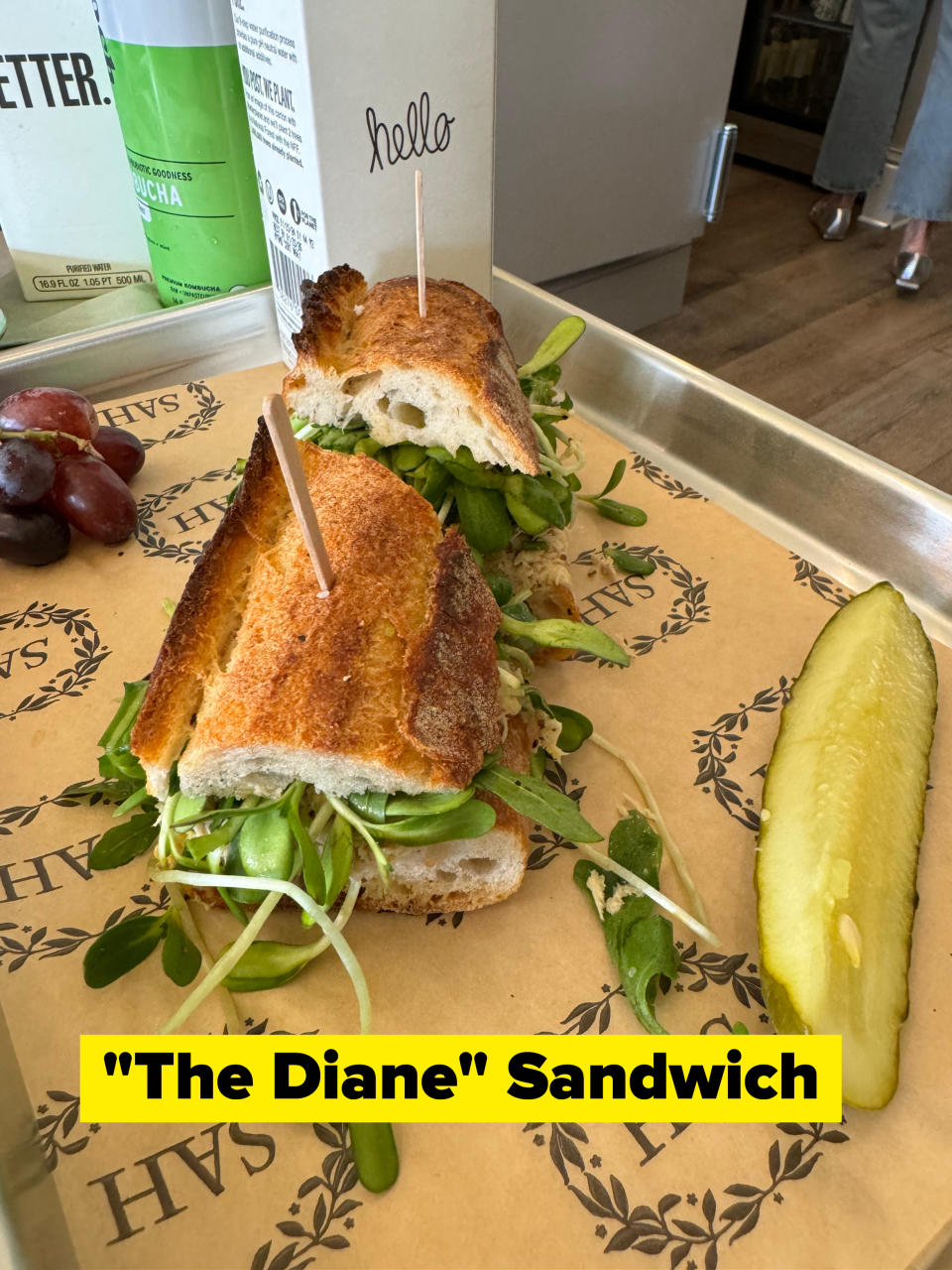 A freshly made baguette sandwich with greens and a pickle slice on a tray. Grapes and a carton of matcha are also visible. The sandwich has a toothpick with a "hello" sign