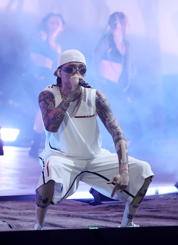 Peso Pluma performing on stage with a microphone, wearing a tank top, shorts, a hat, and sunglasses