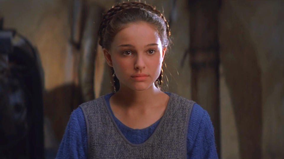 Natalie Portman stands in a workshop with a puzzled look in Star Wars: The Phantom Menace.