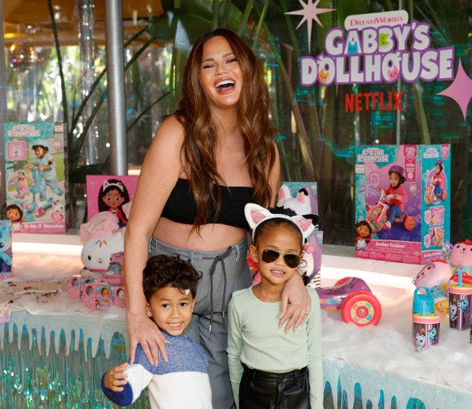 Chrissy laughing as she holds her two eldest children at a Netflix event for Gabby's Dollhouse