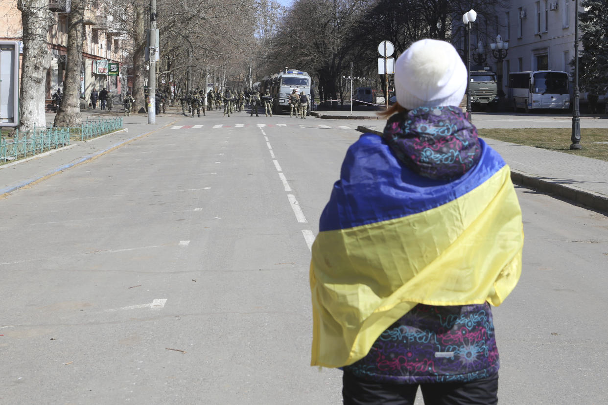 FILE - A woman covered by Ukrainian flag stands in front of Russian troops in a street during a rally against Russian occupation in Kherson, Ukraine, on March 19, 2022. According to Russian state TV, the future of the Ukrainian regions occupied by Moscow's forces is all but decided: Referendums on becoming part of Russia will soon take place there, and the joyful residents who were abandoned by Kyiv will be able to prosper in peace. In reality, the Kremlin appears to be in no rush to seal the deal on Ukraine's southern regions of Kherson and Zaporizhzhia and the eastern provinces of Donetsk and Luhansk. (AP Photo/Olexandr Chornyi)
