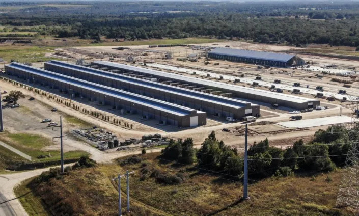 An aerial shot shows rows of long sheds on land surrounded by power lines.