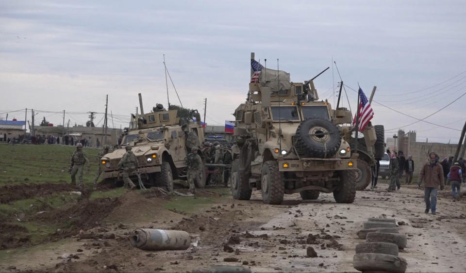 In this frame grab from video, people and soldiers gather next to an American military convoy stuck in the village of Khirbet Ammu, east of Qamishli city, Syria, Wednesday, Feb. 12, 2020. The Syrian official news agency SANA, said Wednesday, that locals had gathered at an army checkpoint, pelting the U.S. convoy with stones and taking down a U.S. flag flying on a vehicle when troops fired with live ammunition and smoke bombs. (AP Photo)