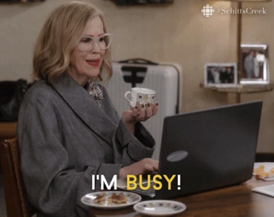 Moira Rose, wearing a gray robe and glasses, sips coffee while working on a laptop. Caption reads "I'M BUSY!"