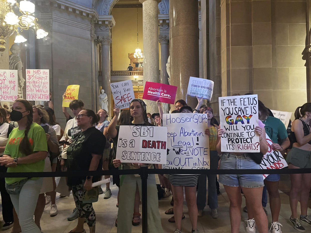 A few dozen protesters are seen standing behind a roped-off area inside a statehouse holding signs reading: If the fetus you save is gay, will you continue to protect its rights? and SB1 is a death sentence, among others.