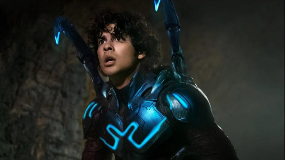 A young Latino man wears a neon blue superhero costume, the blue lit up, with his helmet down, showing his face. There are two antennae sticking up from his shoulders.