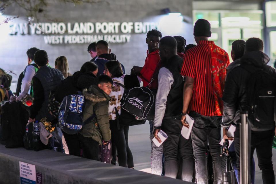 People, some holding white papers, wait outside a building with a sign that says San Ysidro Land Port of Entry