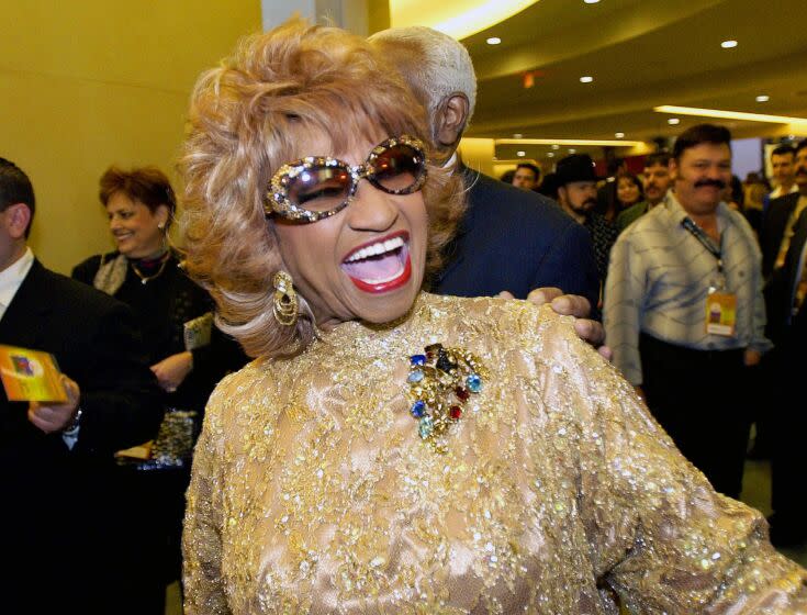 woman, celia cruz, smiling with mouth open, with curled blond hair, in large sunglasses and wearing gold, shimmering dress