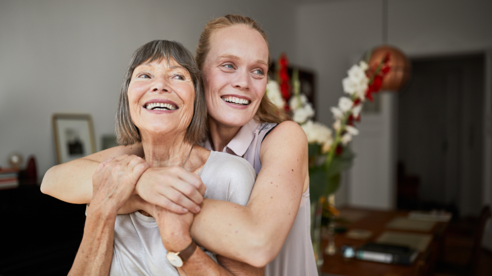 Cheerful mature woman embracing senior mother at home and looking away. 