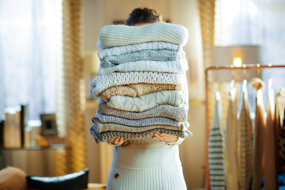 woman in white sweater and skirt in house in sunny winter day holding huge pile of sweaters.