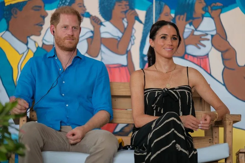 Prince Harry and Meghan Markle sit next to each other on a bench, smiling. Harry is wearing a blue shirt and Meghan a black dress.