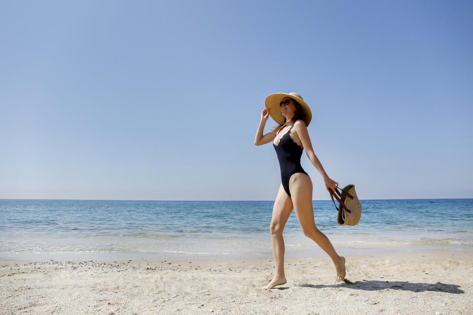 Young beautiful woman wearing one piece swimsuit, broad brim straw hat posing with a beach bag. Female fashion model at sandy beach on beautiful sunny day. Mediterranean sea background. Copy space