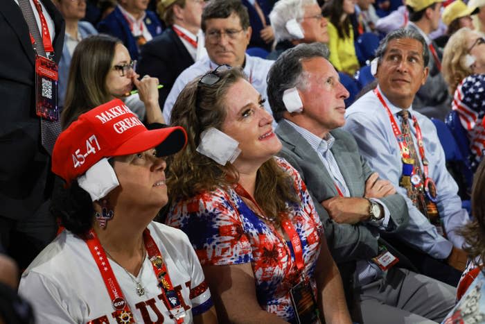 Menschen bei einer Kundgebung von Donald Trump, einige mit Gehörschutz und patriotischer Kleidung, sitzen da und hören aufmerksam zu