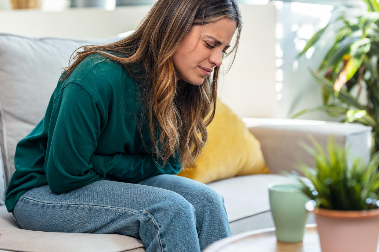 Shot of unhealthy young woman with stomachache leaning on the couch in the living room at home.