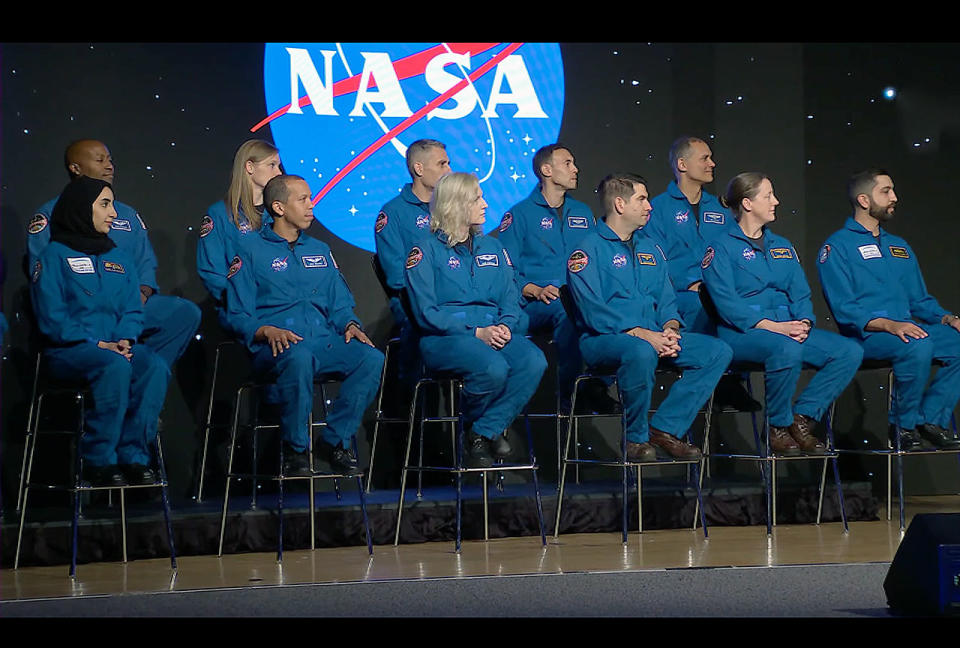 11 people in blue flight suits sit on a stage with a projection of the NASA logo on the wall behind them