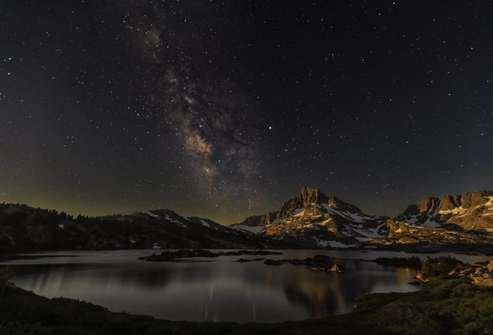 A lake beneath snow-flecked mountains, with many stars visible in the night sky.