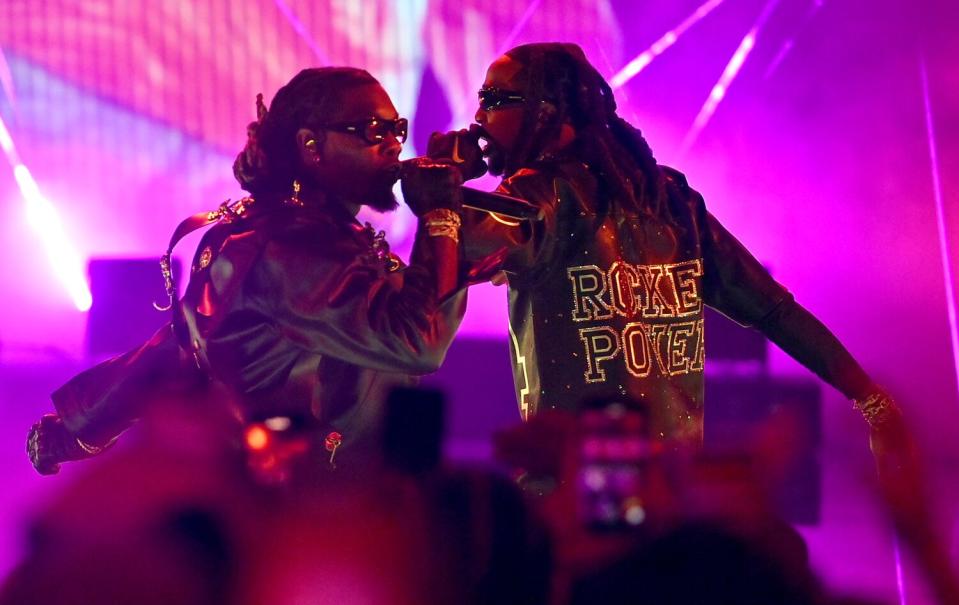 Offset and Quavo rapping into microphones, holding it to their mouths on stage, wearing black jackets and sunglasses