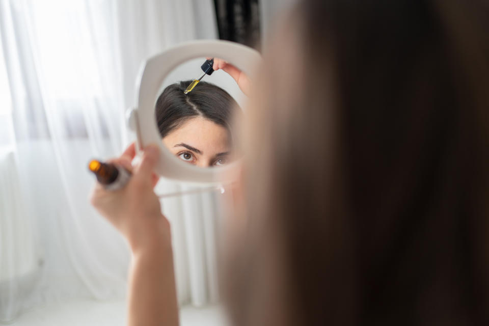 Woman applying oil to scalp in mirror.
