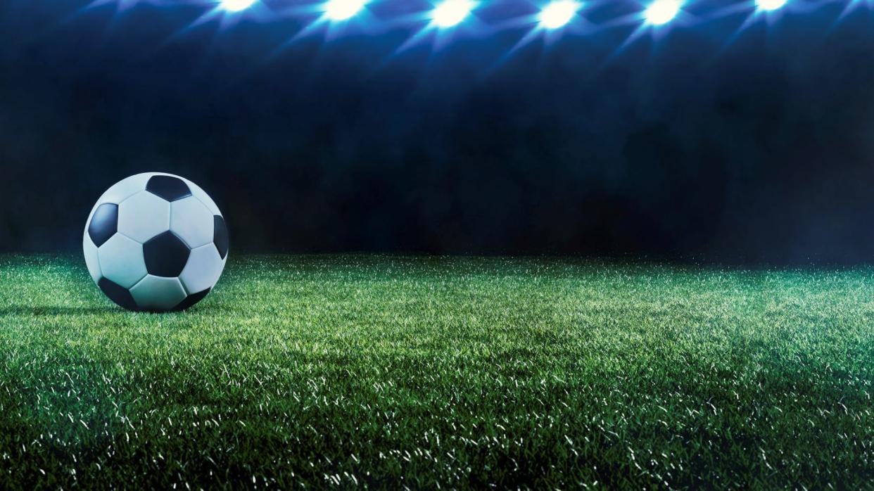 Football or soccer background with a row of spotlights illuminating a ball on the green turf in a stadium in a sports championship or World Cup event in a panorama banner.