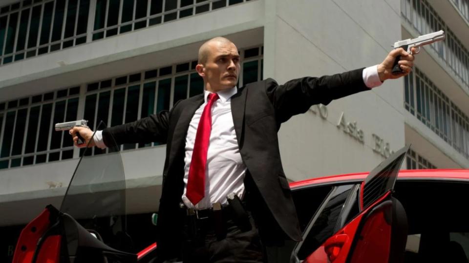 A man in a suit with a red tie stretches his arms out and aims guns in either direction in front of a red sports car